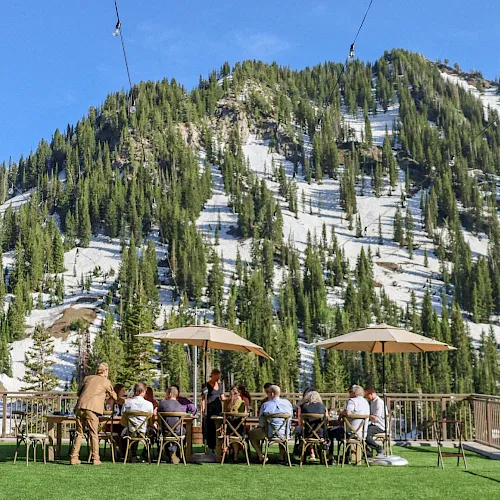A sunny ski resort scene with chairlift cables over snowy mountains, tall pines, and people dining outdoors under umbrellas.
