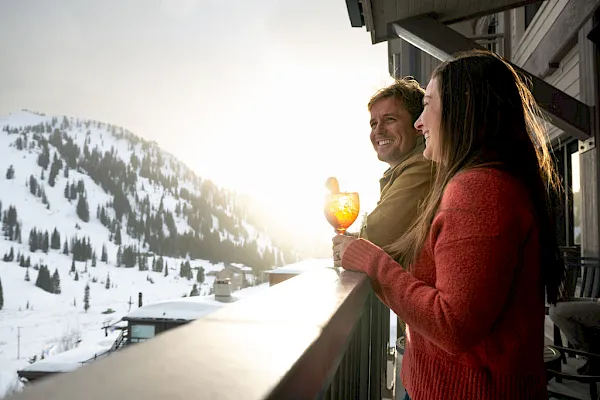 A couple smiles on a snowy mountain balcony, toasting with warm drinks as winter sun lights the slopes.