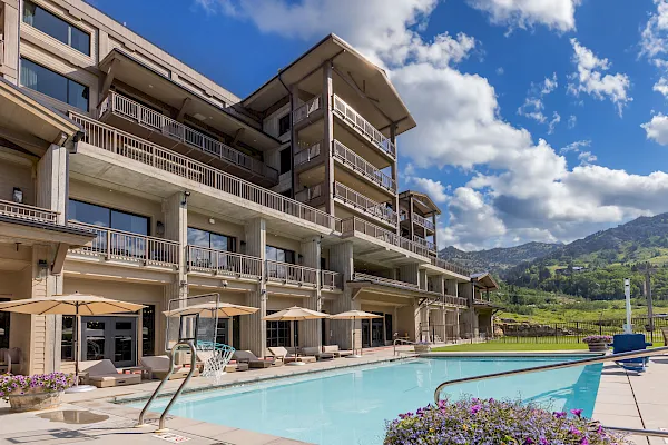 A sunny hotel with multiple balcony floors overlooks a swimming pool and a lush green mountain valley.