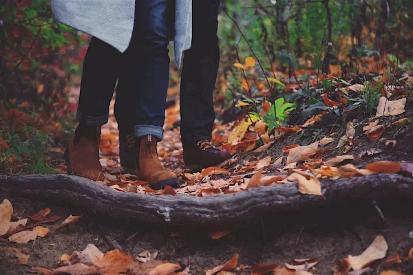 A person’s legs in jeans and brown boots walk through a forest floor scattered with colorful autumn leaves, embracing fall.
