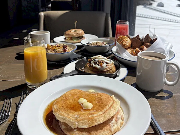 A breakfast spread with pancakes, a savory potato dish, eggs/benedict, pastries, orange juice, and coffee on a table.