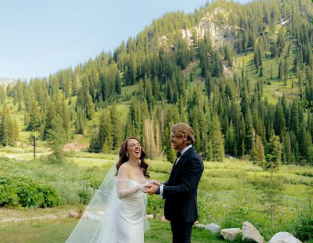 A bride and groom in wedding attire laugh outdoors in a green meadow with a pine-covered mountain and blue sky behind them.