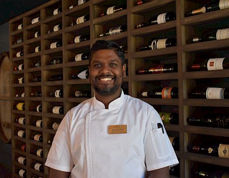 A smiling chef in a wine cellar, standing in front of shelves filled with neatly arranged wine bottles, in a warm setting.