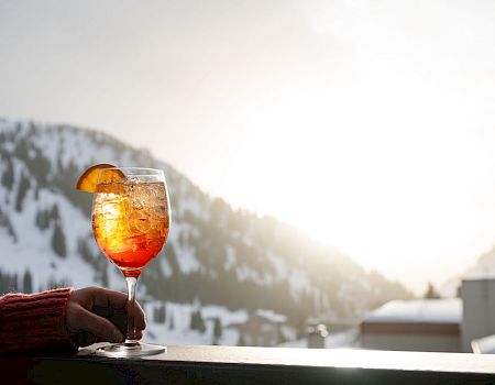 A hand holds an orange-garnished cocktail in a glass, with snowy mountain scenery in the background at sunset.