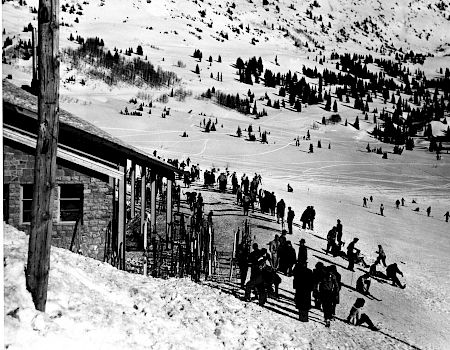Black-and-white photo of skiers and spectators on a snowy slope near a lodge, with tracks and pine forests in the distance.