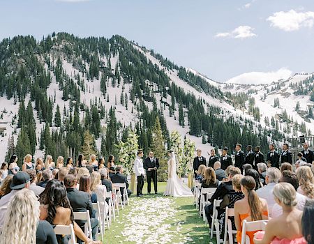 An outdoor wedding ceremony in snowy mountains: the bride and groom stand before a group of attendants while guests watch from chairs.