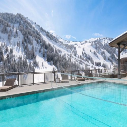 An outdoor heated pool on a chalet terrace with lounge chairs, set against snowy alpine mountains under a clear blue sky.
