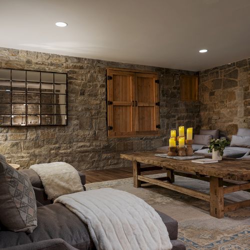 A cozy stone-walled living room with sofas, a rustic coffee table, wooden doors, candles, and wall art.