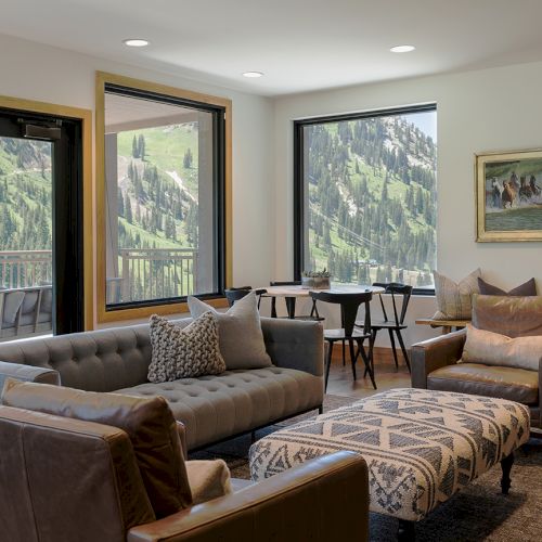 A cozy living room with a sofa and armchairs, framed by large windows showing lush green mountains.