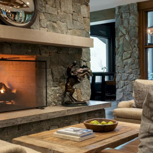 A cozy stone fireplace and lounge area with sofas, a wooden coffee table, and warm lighting through large windows, always.