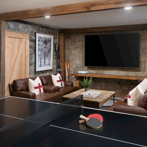 A modern lounge with a ping-pong table, brown leather sofas, stone walls, framed art, a TV, and a wood mantle fireplace.