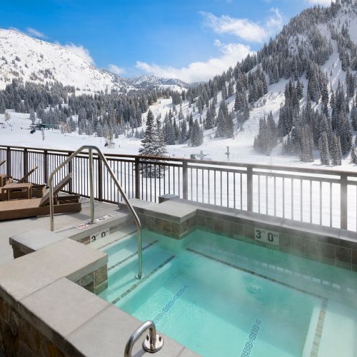 An outdoor hot tub with steps and lounge seating overlooks a snowy ski valley of pine trees and mountains under a blue sky.