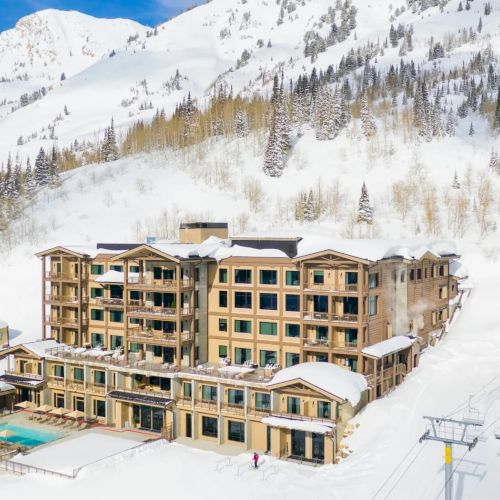A snowy mountain ski resort with a large hotel, ski chairlifts, and a backdrop of pine-covered alpine slopes under clear blue skies.