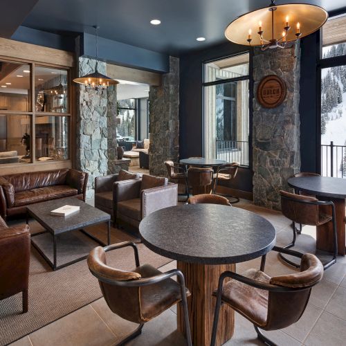 Rustic lodge lounge with leather sofas, round tables, stone columns, pendant chandeliers, and large windows showing snowy pine trees outside.
