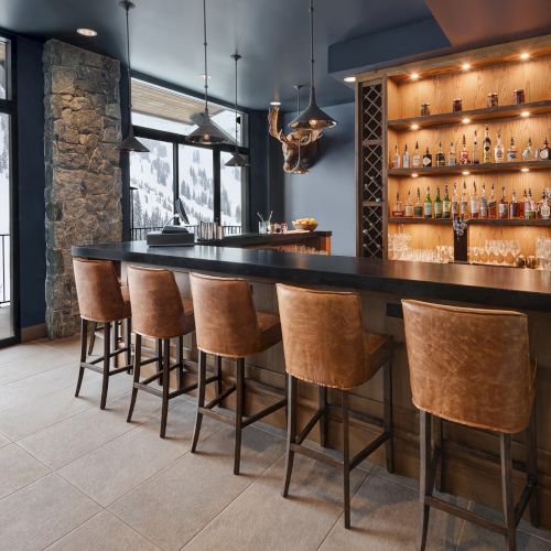 Cozy mountain bar with leather stools, stone column, ski-view windows, and a wall liquor display with lit bottles.