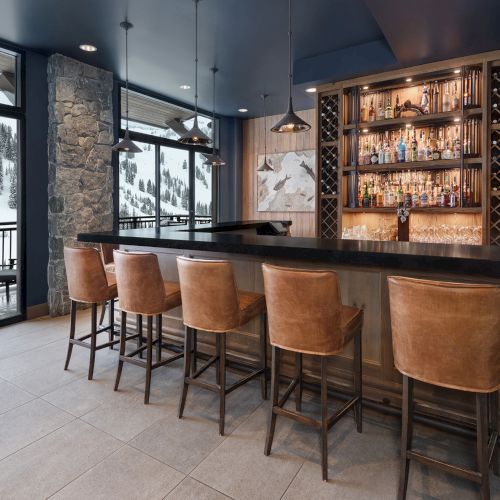 A cozy lodge-style bar with leather stools, hanging lights, stocked liquor shelves, and snowy mountain views through large windows.
