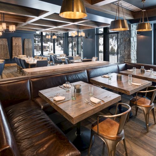 A cozy alpine restaurant dining room with wooden tables, leather booths, and hanging copper pendant lights, overlooking snowy mountains.