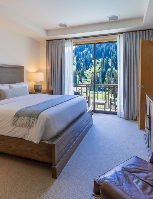 A modern hotel bedroom with a king bed, TV, and balcony doors showing pine-covered mountains.