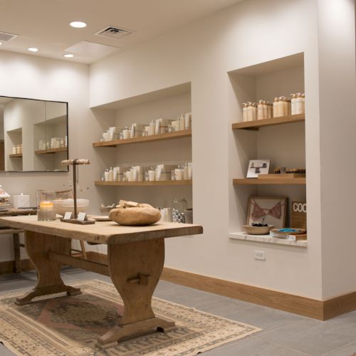 A bright spa shop with shelves of skincare and candles, a wooden treatment table, rugs, and mirrors.