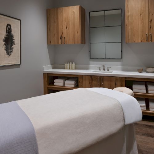 A modern spa bathroom with a massage table, wooden vanity and mirror, twin sinks, neatly folded towels, and minimalist decor.