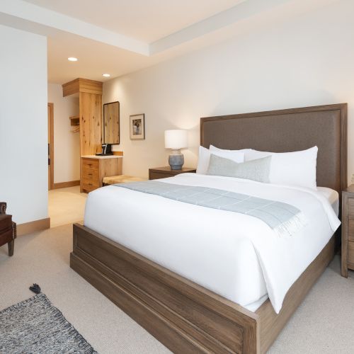 A bright modern bedroom with a large upholstered bed, bedside lamps, wood cabinets, and a small seating area with artwork.