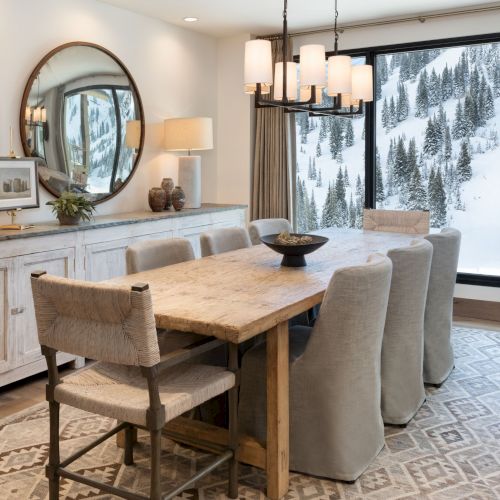 A bright modern dining room with a rustic wooden table, cushioned chairs, pendant lights, and floor-to-ceiling windows showing snowy mountains.