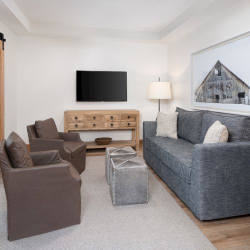 A bright living room with a flat-screen TV, wood media console, gray and brown sofas, coffee ottoman, and a large framed barn print.
