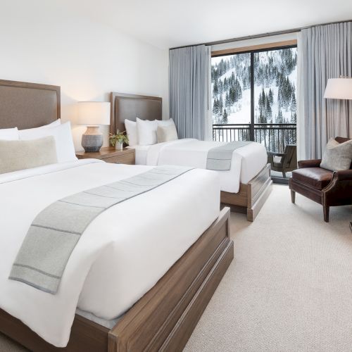 A modern hotel bedroom with two made-up beds, gray curtains, a mountain forest view, bedside lamps, and a flat-screen TV.