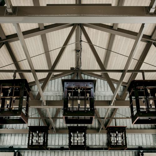 A high-ceiling warehouse ceiling with steel trusses and three hanging bottle-filled crates suspended by chains, inside an industrial bottling line.