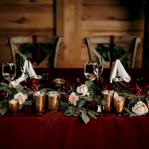 An elegant banquet table set with wine glasses, golden goblets, candles, and red-and-white roses on a dark red cloth.