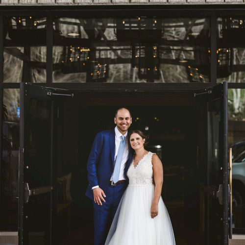 A smiling bride and groom pose at an open building entrance, seen through glass with warm lights inside.