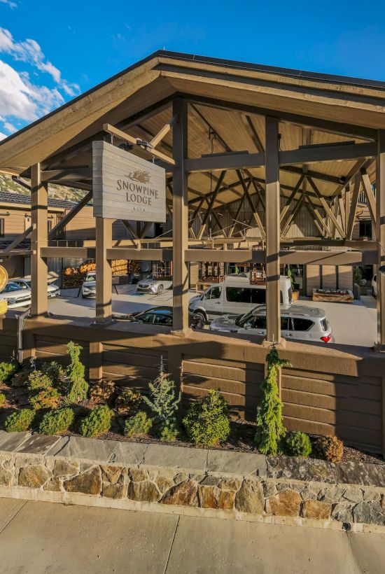 Snowpine Lodge at Alta sits under a wooden porte-coch&egrave;re with cars parked nearby and mountain views under a blue sky.
