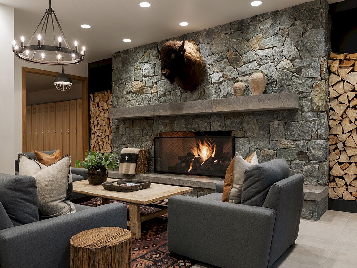 Cozy living room with a stone fireplace, mounted bison head, gray sofas, wooden coffee table, and warm chandelier lights.