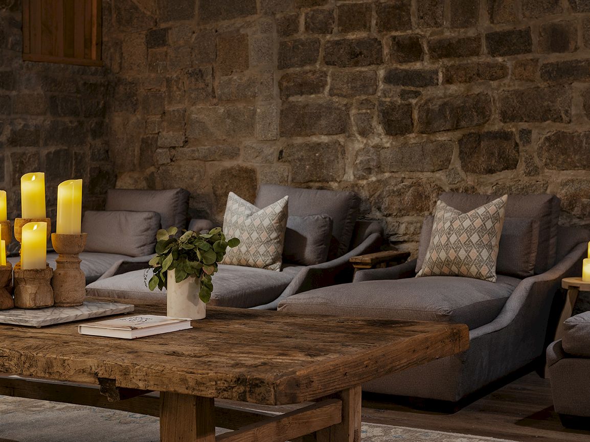Cozy living room with a gray sectional sofa, patterned pillows, a rustic wooden coffee table, and lit candles by the stone wall.