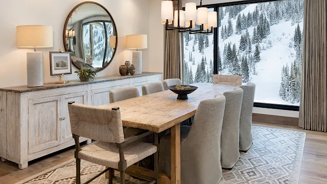 A cozy dining room with a rustic wooden table, upholstered chairs, pendant lights, and large windows showing snowy pine trees outside.