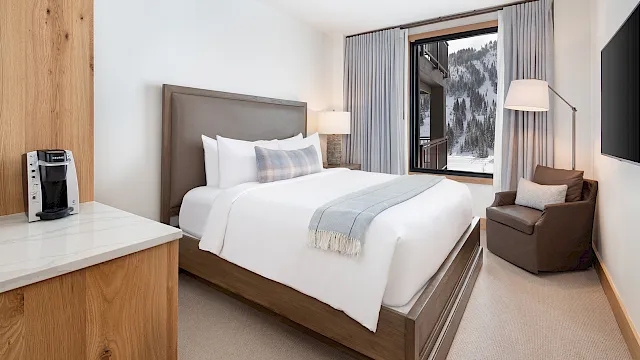 Modern hotel room with a neatly made bed, gray curtains, bedside lamps, a window to snowy mountains, and a Keurig coffee maker.