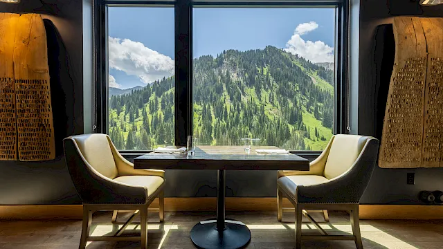 A cozy seating area with two chairs and a small table, gazing at a lush mountain valley through large windows on a sunny day.