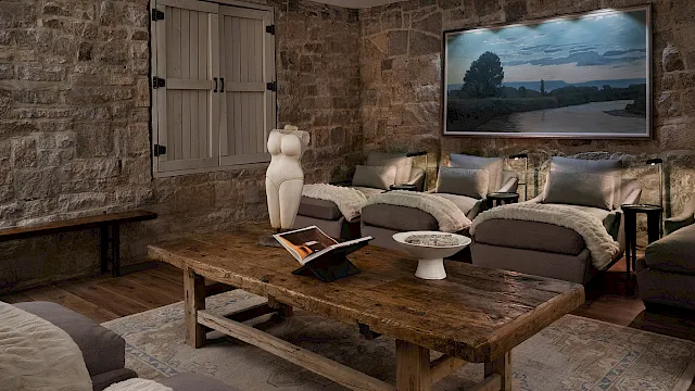A rustic living room with stone walls, shuttered window, sofas, a wooden coffee table, wall art, lamps, and a small sculpture.