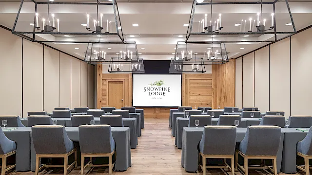 A conference hall with rows of gray chairs, hanging chandeliers, and a stage screen displaying &ldquo;Snowpine Lodge,&rdquo; ready for meetings.