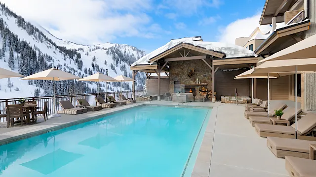 A snowy mountain resort patio with a turquoise outdoor pool, lounge chairs, umbrellas, and a stone fireplace beside the ski slopes.