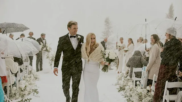A snowy outdoor wedding: a groom and bride walk down the aisle as guests hold umbrellas.