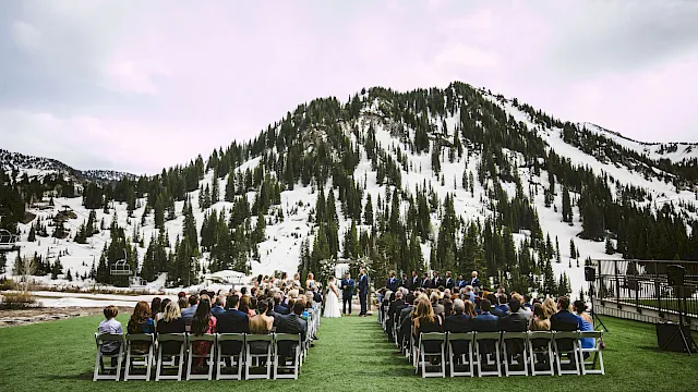 Outdoor wedding ceremony on a green lawn with guests seated, a couple at the aisle, and snowy mountain peaks behind.