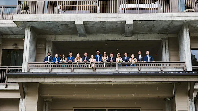A wedding party poses on an outdoor balcony in blue suits and dresses holding bouquets.