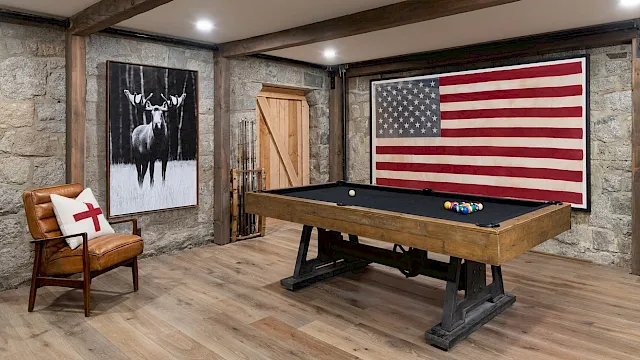 Rustic game room with a wooden pool table, deer wall art, and a large American flag backdrop.