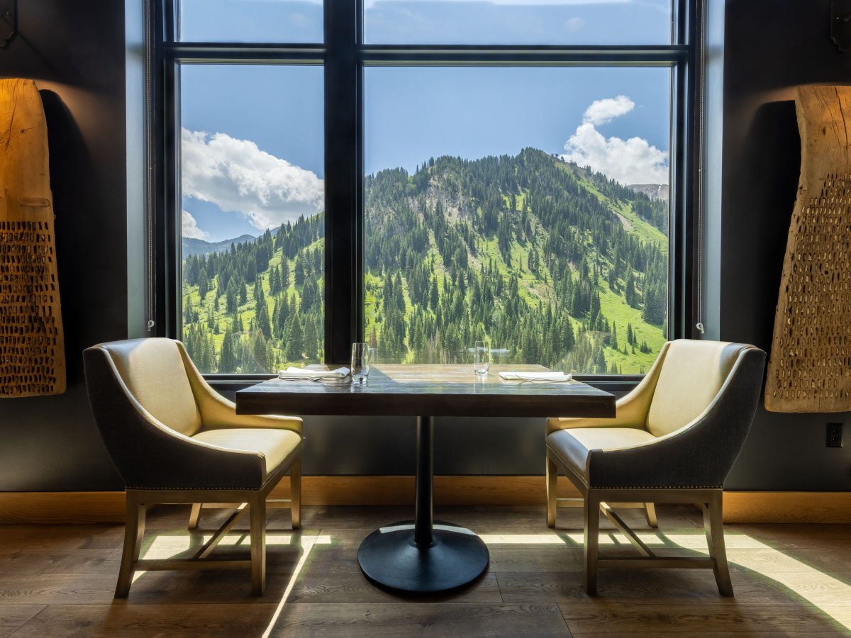 A cozy caf&eacute; table with two chairs by big windows, framing lush green mountains and clouds outside.