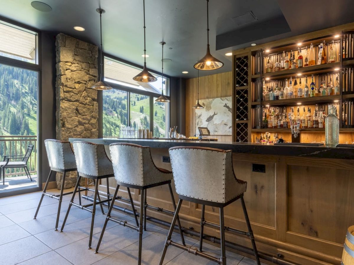 A cozy mountain bar with a long countertop, tall stools, hanging lights, and shelves of liquor bottles overlooking the scenic hills.