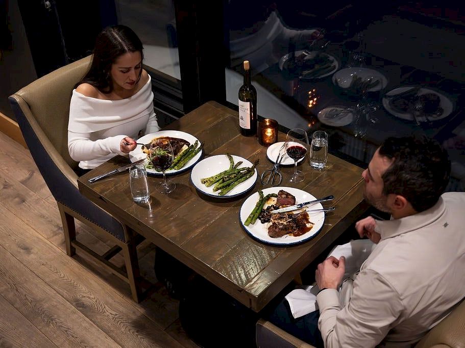 A couple in white outfits dine at a restaurant table with plated meals, asparagus, and a bottle of wine.