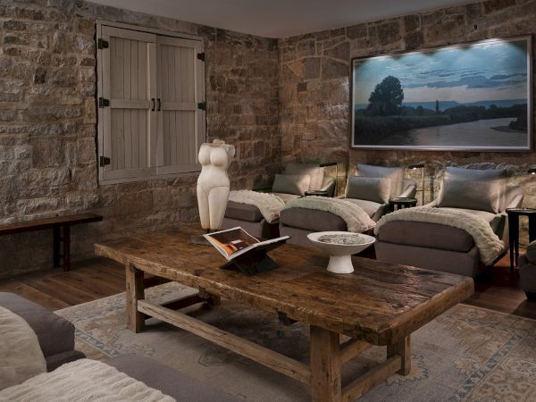 Rustic stone living room with plush sofas, wooden coffee table, wall art landscape, and shuttered window, cozy lighting.