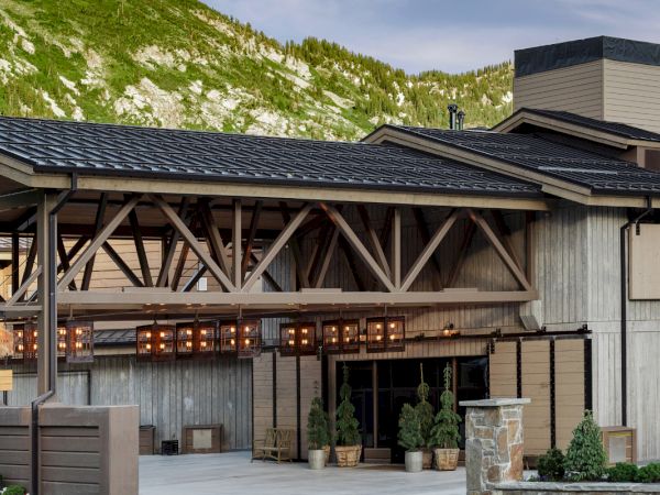 A timber lodge called Snowpine Lodge with lit windows sits in a green mountain valley with snow-dotted hills behind it.