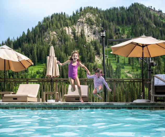 A woman and a little girl leap into a swimming pool in summer, splashing while overlooking green mountains.